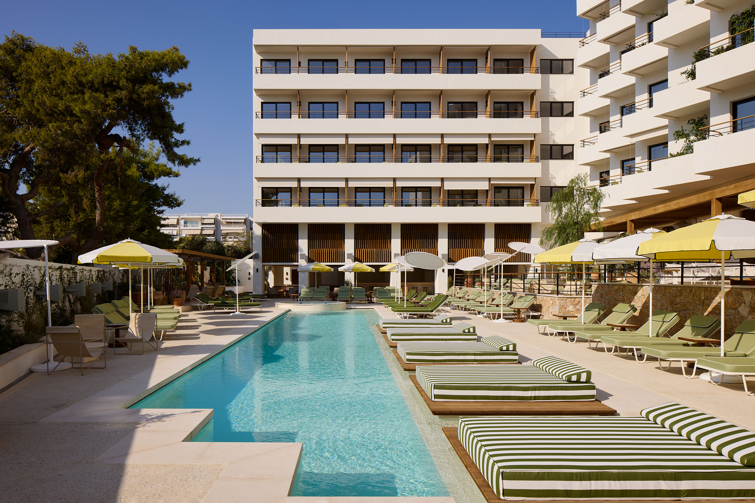 Outdoor pool area at Ace Hotel & Swim Club Athens, featuring striped green and white lounge beds, yellow and white umbrellas, and a modern white facade with balconies overlooking the pool. The poolside is lined with lush greenery and stylish seating, creating a relaxed and inviting atmosphere.